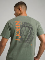 Person wearing a green T-shirt with a dark lion illustration, orange “1 John 4:4” and cross, and the verse “You, dear children, are from God and have overcome them, because the one who is in you is greater than the one who is in the world.”