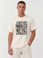 A man wearing a cream-colored t-shirt with a black graphic print that reads 'JESUS IS KING' alongside an illustration of a lion, paired with light beige pants, against a plain light gray background.