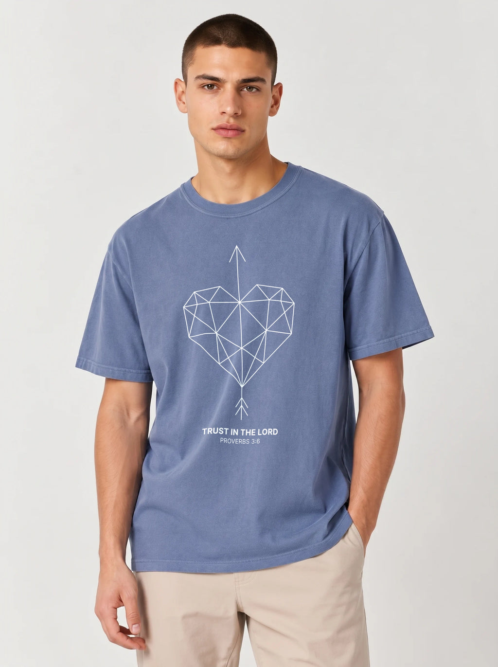 A man wearing a heathered blue t-shirt with the a geometric heart pierced by an upward-pointing arrow, paired with the verse Proverbs 3:6: "Trust in the Lord.", paired with beige trousers, against a light gray studio background.