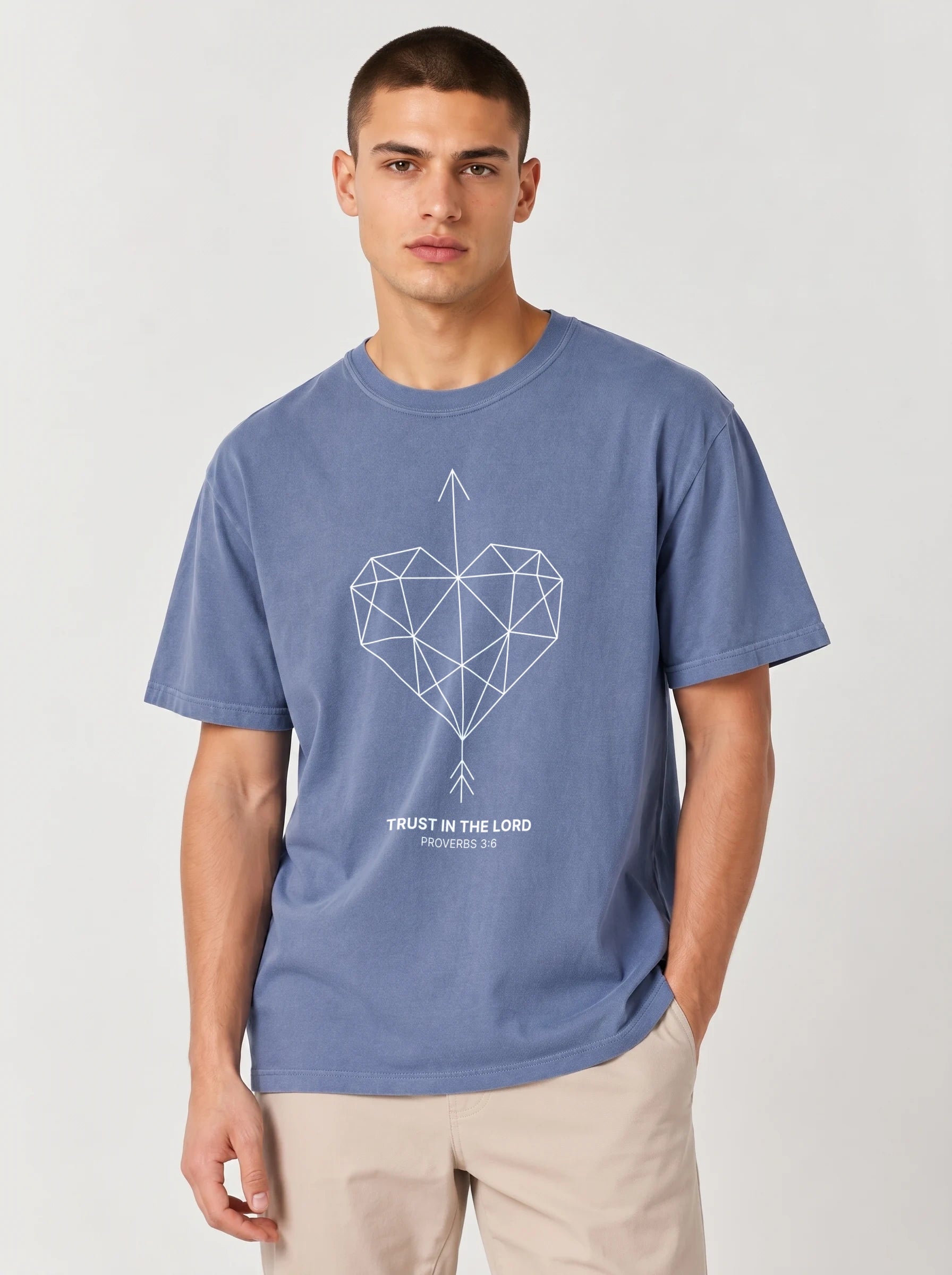 A man wearing a heathered blue t-shirt with the a geometric heart pierced by an upward-pointing arrow, paired with the verse Proverbs 3:6: "Trust in the Lord.", paired with beige trousers, against a light gray studio background.