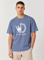 A man wearing a faded blue crewneck t-shirt with a white graphic of a crown of thorns and the text 'LORD & SAVIOR' on the front, paired with beige pants, against a light gray studio background.