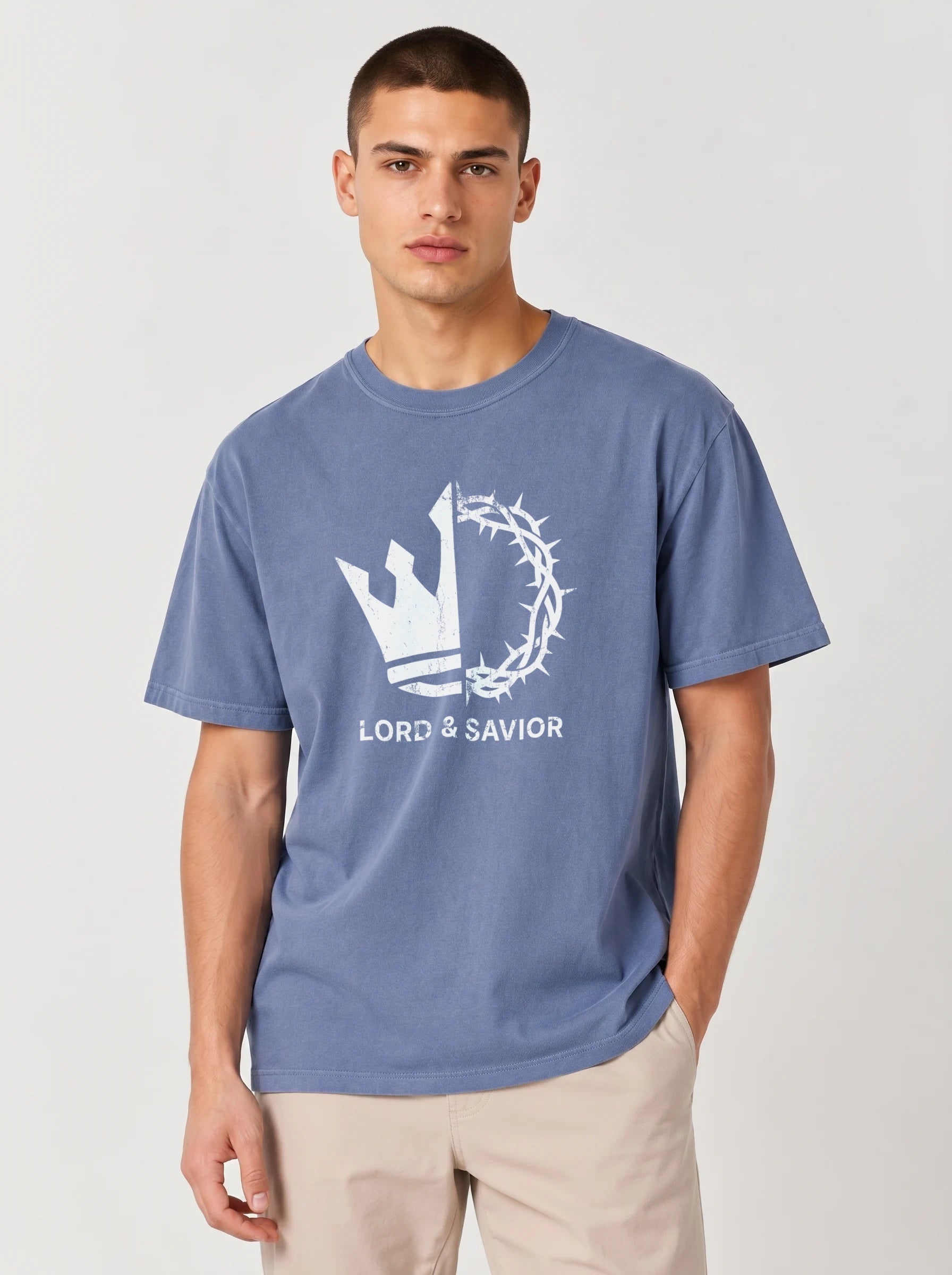 A man wearing a faded blue crewneck t-shirt with a white graphic of a crown of thorns and the text 'LORD & SAVIOR' on the front, paired with beige pants, against a light gray studio background.