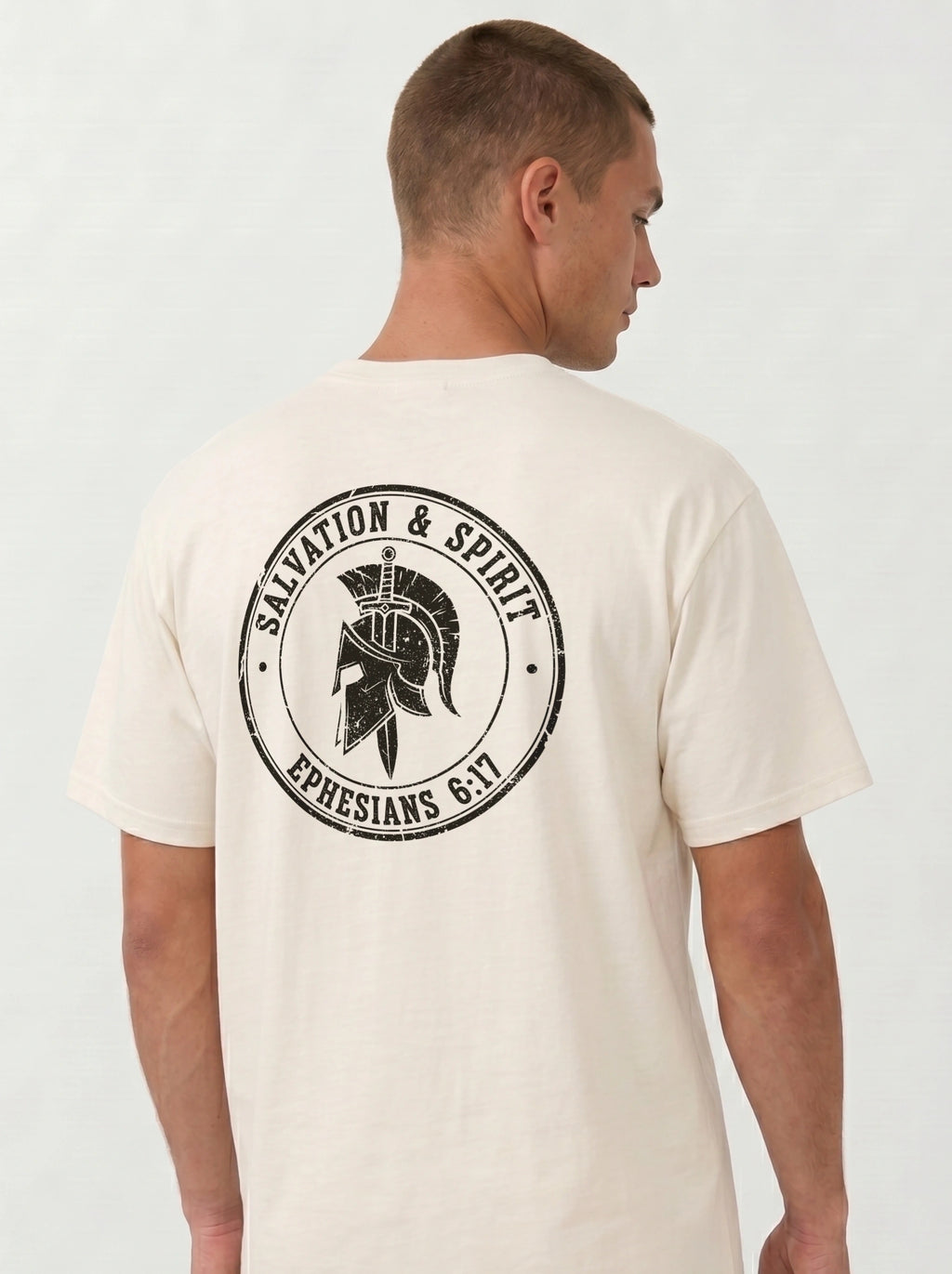 A man viewed from the back wearing a cream-colored t-shirt with a large black distressed graphic on the back featuring a Spartan helmet and the text 'SALVATION & SPIRIT' and 'EPHESIANS 6:17'.