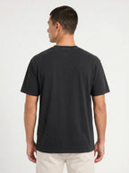 A man viewed from the back wearing a plain black short-sleeve crewneck t-shirt and light beige pants, set against a clean white studio background.