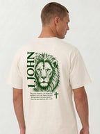 Person wearing a cream T-shirt with a green lion illustration, cross, “1 John 4:4,” and the verse text, “You, dear children, are from God and have overcome them, because the one who is in you is greater than the one who is in the world.”