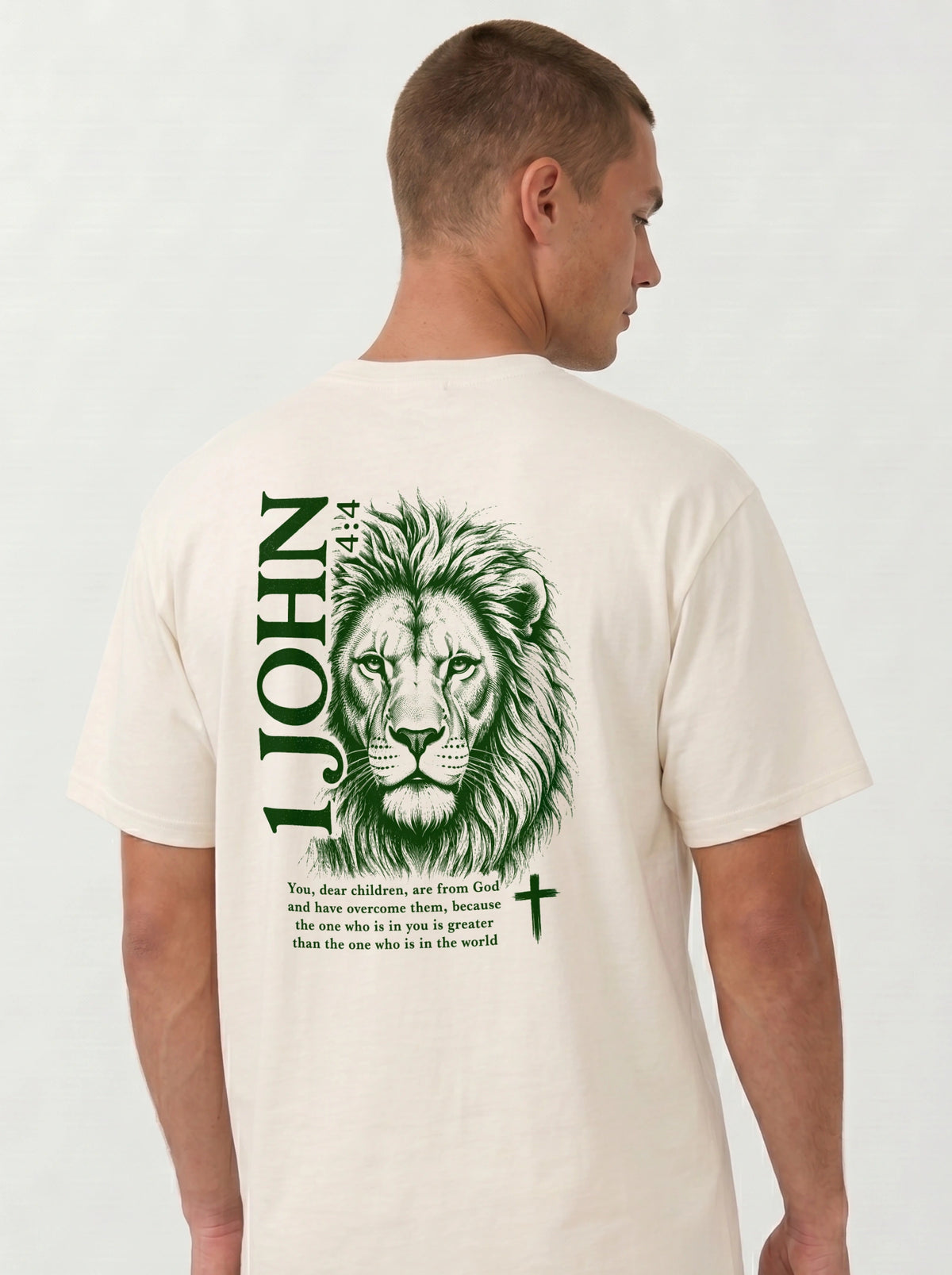 Person wearing a cream T-shirt with a green lion illustration, cross, “1 John 4:4,” and the verse text, “You, dear children, are from God and have overcome them, because the one who is in you is greater than the one who is in the world.”