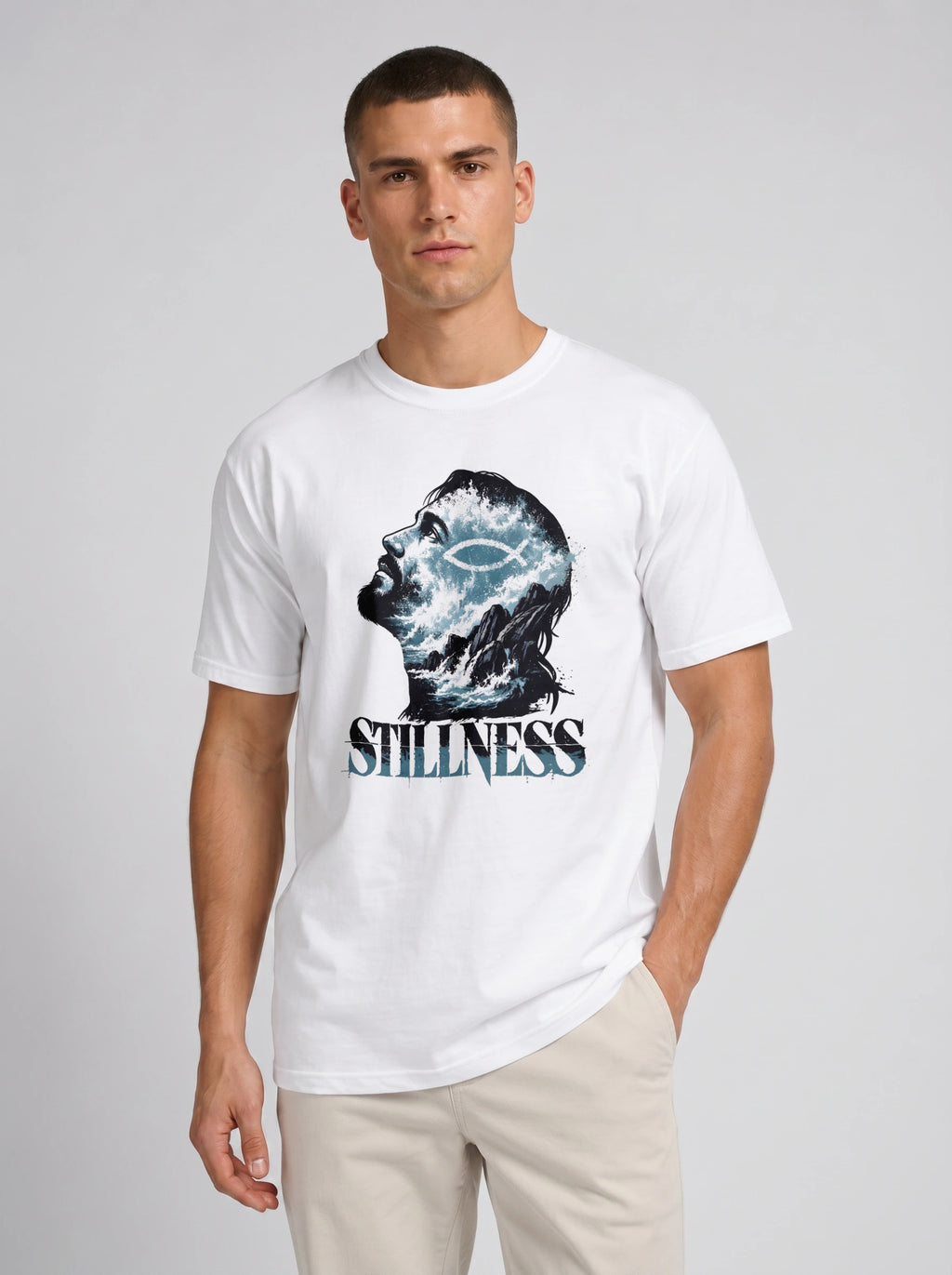 A man wearing a white crew-neck t-shirt with a graphic print of a profile head filled with blue and white waves, and the word 'STILLNESS' printed below in a distressed blue font. He is also wearing beige pants, standing against a light gray studio background.