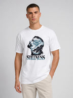 A man wearing a white crew-neck t-shirt with a graphic print of a profile head filled with blue and white waves, and the word 'STILLNESS' printed below in a distressed blue font. He is also wearing beige pants, standing against a light gray studio background.