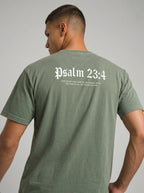 A man wearing an olive green t-shirt, viewed from the back. The shirt features the text 'Psalm 23:4' printed in a white gothic font, with a smaller bible verse underneath, set against a plain light gray background.