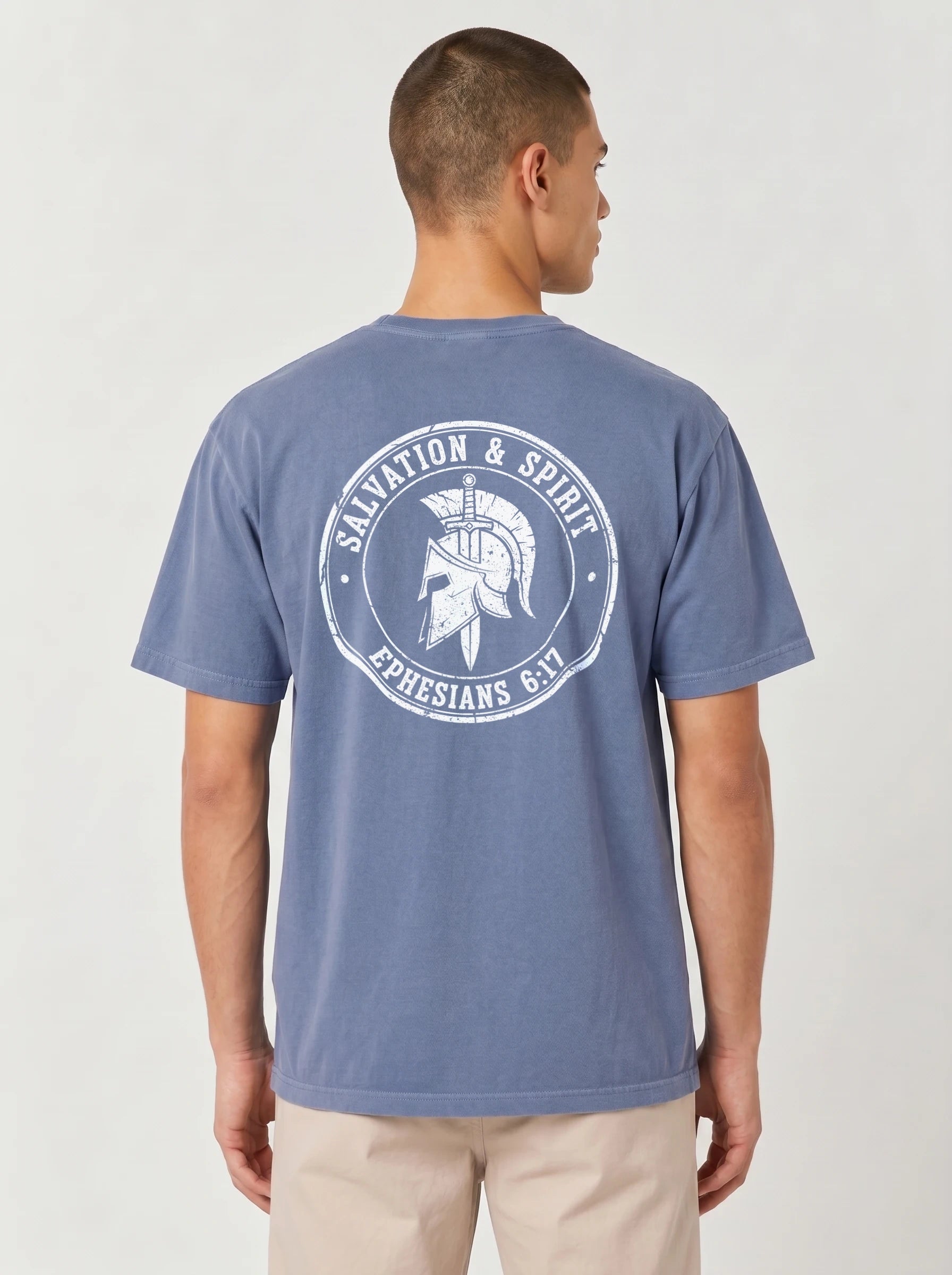 A man viewed from the back wearing a slate blue crewneck t-shirt with a distressed white graphic on the back that reads 'Salvation & Spirit Ephesians 6:17' around a Spartan helmet. He is also wearing beige pants, set against a plain light gray studio background.