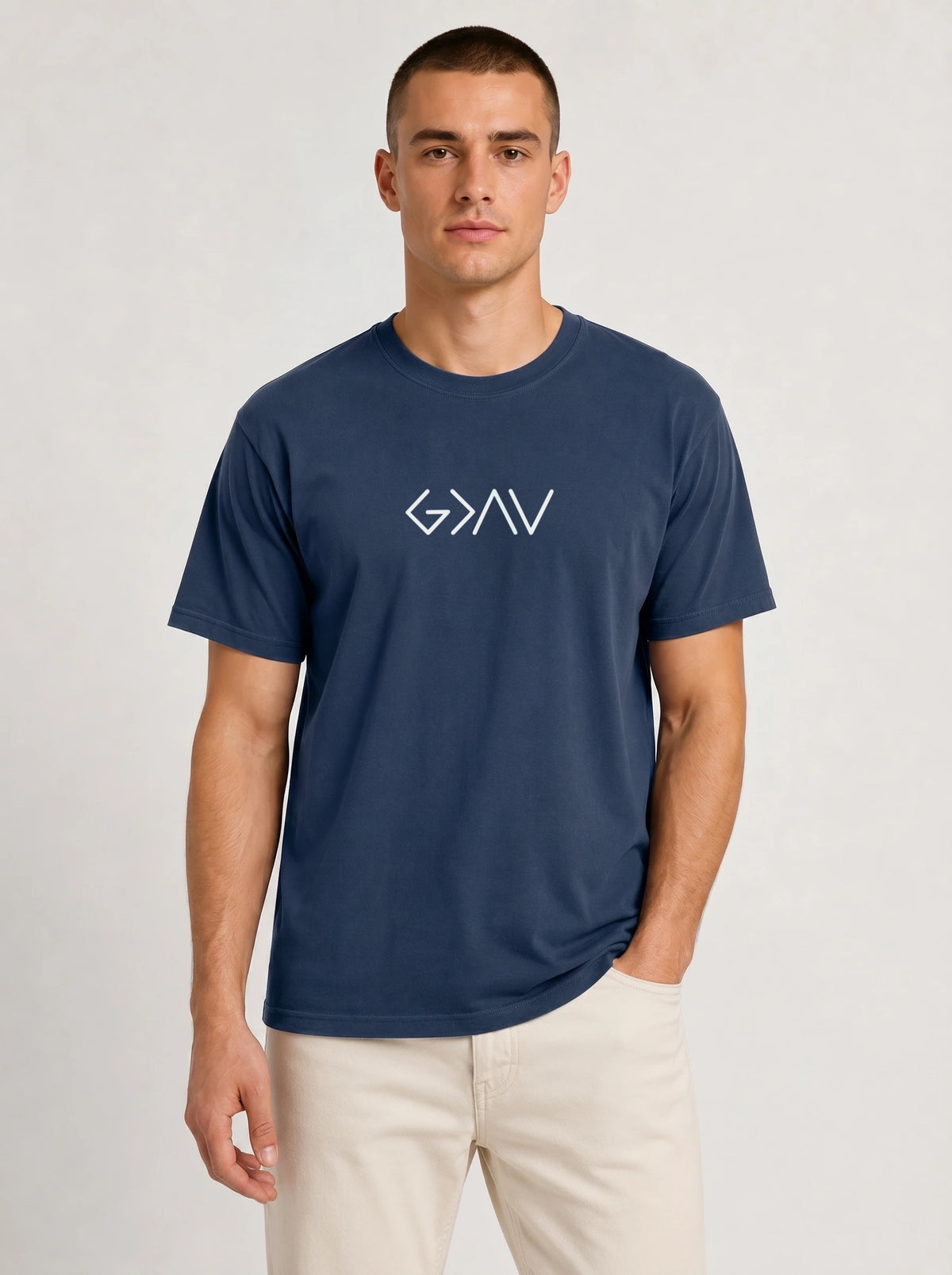 A man wearing a navy blue crewneck t-shirt with the white printed text 'God Is Greater Than Highs and Lows', paired with light beige pants, against a light gray studio background.