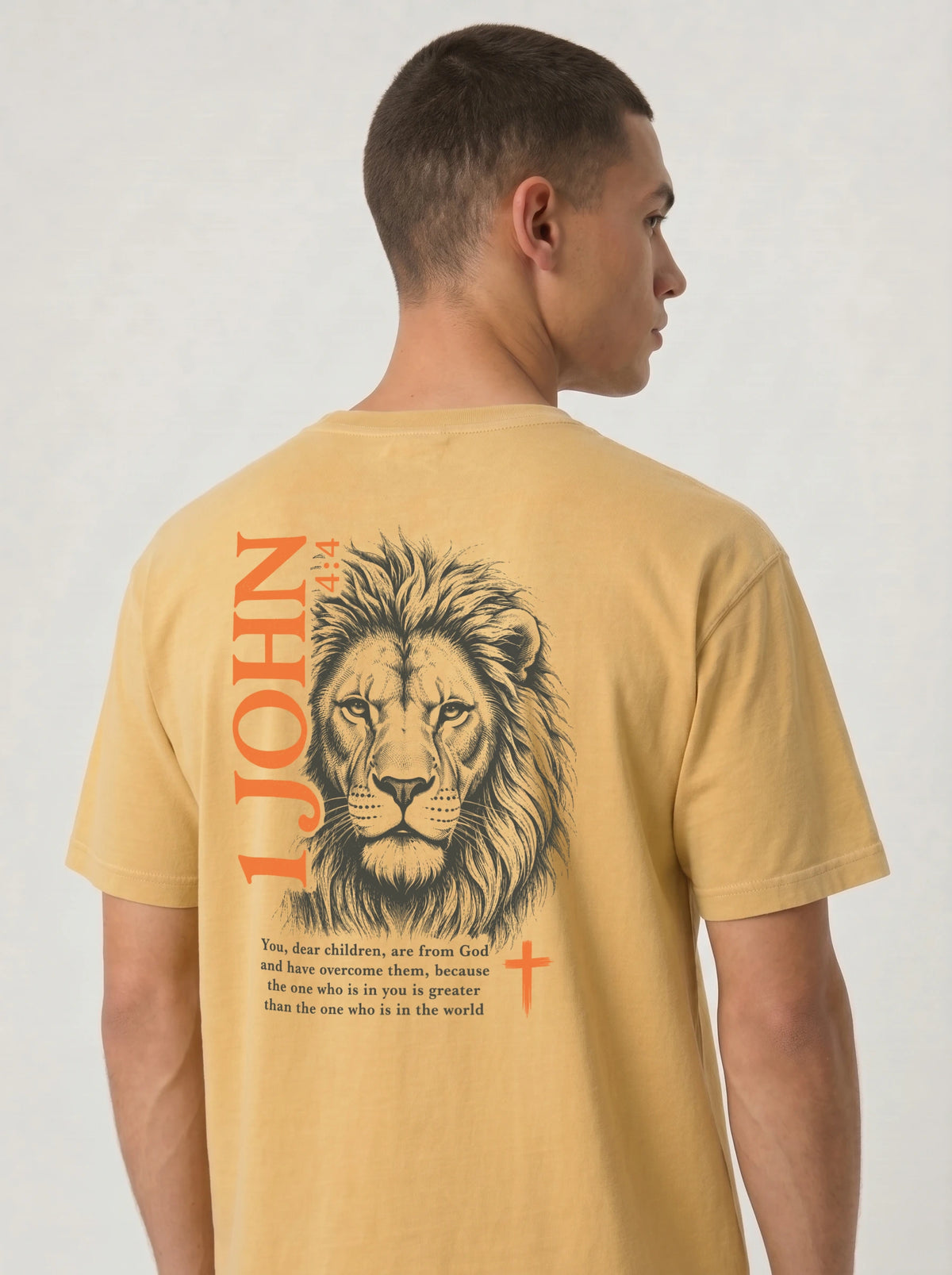 Person wearing a yellow T-shirt with a dark lion illustration, orange “1 John 4:4” and cross, and the verse “You, dear children, are from God and have overcome them, because the one who is in you is greater than the one who is in the world.”