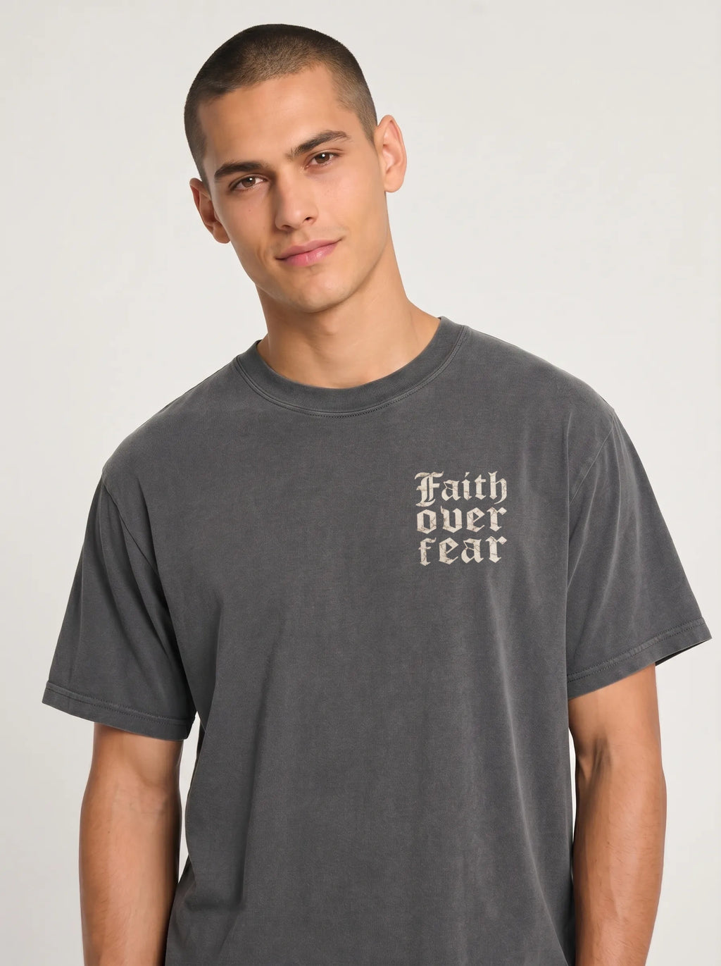 A man wearing a dark gray washed t-shirt with the text 'Faith over fear' printed in a sand colored Gothic font on the left chest, photographed against a light gray studio background.