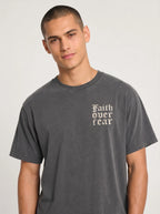 A man wearing a dark gray washed t-shirt with the text 'Faith over fear' printed in a sand colored Gothic font on the left chest, photographed against a light gray studio background.