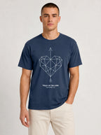 A man wearing a navy blue crewneck t-shirt with a white geometric heart pierced by an upward-pointing arrow, paired with the verse Proverbs 3:6: "Trust in the Lord.", paired with beige pants, against a light gray studio background.
