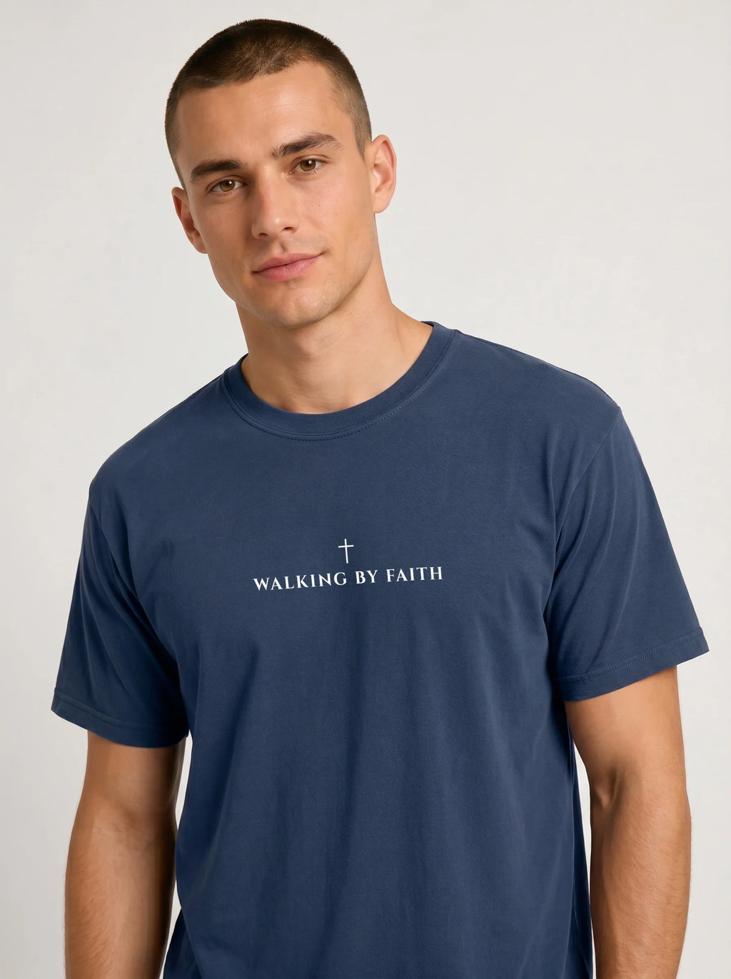 A man wearing a navy blue crewneck t-shirt with a white graphic print on the chest that reads 'Walking By Faith' with a cross above the text