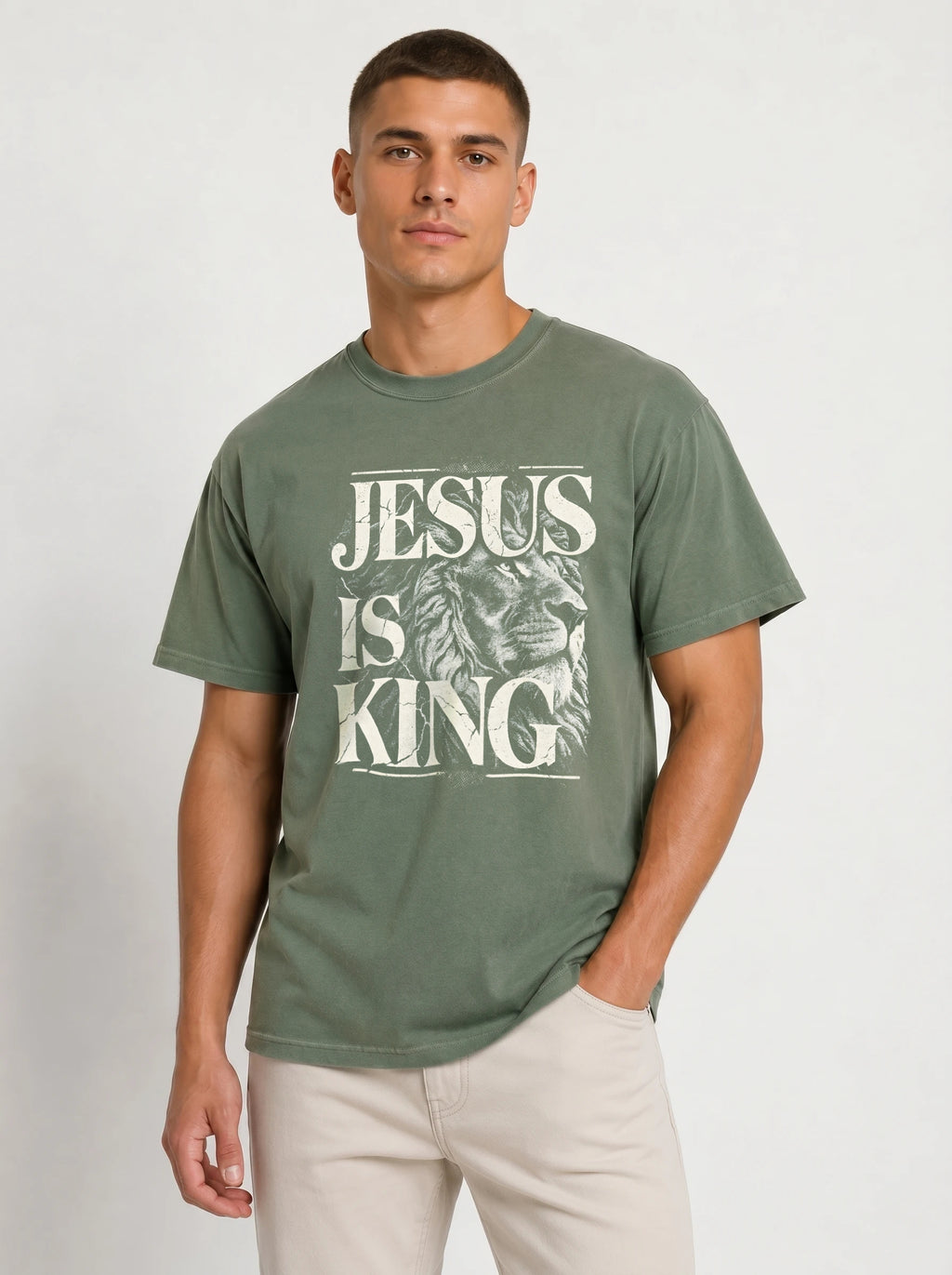 A man wearing a faded olive green t-shirt with a white graphic print that reads 'JESUS IS KING' over a lion illustration, paired with light beige pants, against a plain light gray background.