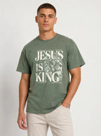 A man wearing a faded olive green t-shirt with a white graphic print that reads 'JESUS IS KING' over a lion illustration, paired with light beige pants, against a plain light gray background.