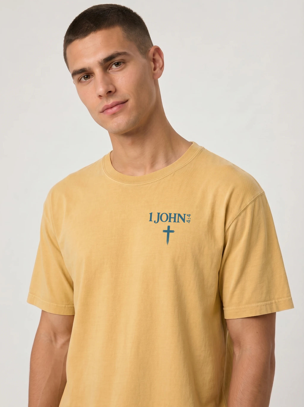 A man wearing a mustard yellow garment-dyed t-shirt, viewed from the front. The shirt has a small chest print reading '1 JOHN 4:4' with a cross symbol beneath it.