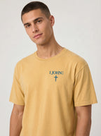 A man wearing a mustard yellow garment-dyed t-shirt, viewed from the front. The shirt has a small chest print reading '1 JOHN 4:4' with a cross symbol beneath it.