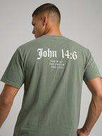 A man wearing an olive green t-shirt, viewed from the back. The shirt features the text 'John 14:6 THE WAY THE TRUTH THE LIFE' printed in white gothic and serif fonts on the upper back.