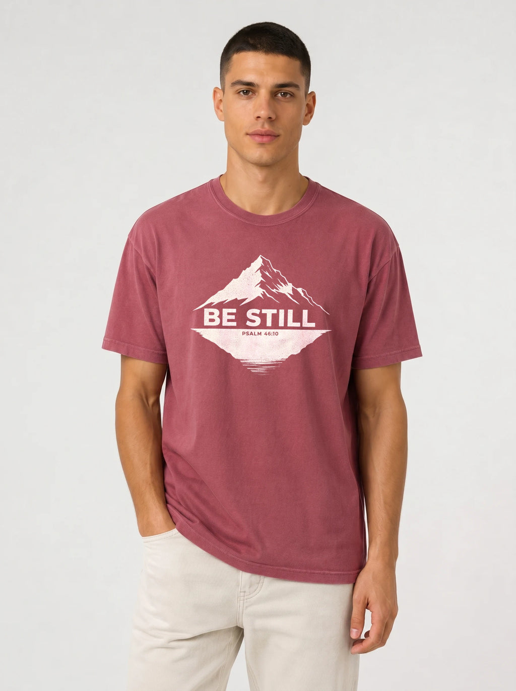 A man wearing a heathered dusty rose t-shirt with a white graphic print of a mountain and the text 'BE STILL' and 'PSALM 46:10' printed on the chest.
