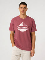 A man wearing a heathered dusty rose t-shirt with a white graphic print of a mountain and the text 'BE STILL' and 'PSALM 46:10' printed on the chest.