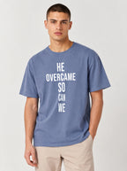 Person wearing a faded blue T-shirt with large white text reading “HE OVERCAME SO CAN WE.”
