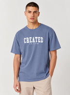 A man wearing a dusty blue crewneck t-shirt with 'CREATED With A Purpose' printed in white across the chest, paired with beige pants, against a light gray studio background.
