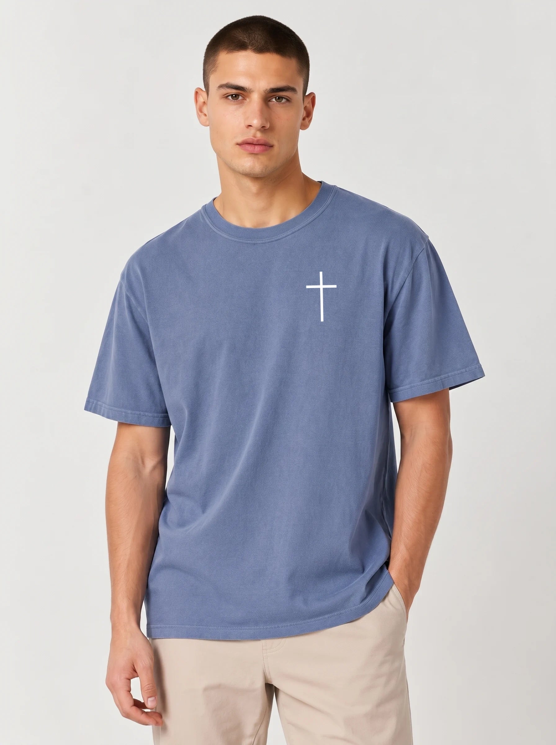 A man wearing a dusty blue crewneck t-shirt with a small white cross printed on the left chest, paired with beige pants, against a light gray studio background.