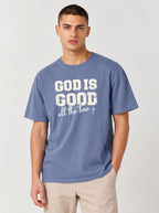 A man wearing a slate blue t-shirt with a white graphic print that reads 'GOD IS GOOD all the time +', paired with beige pants, against a light gray studio background.