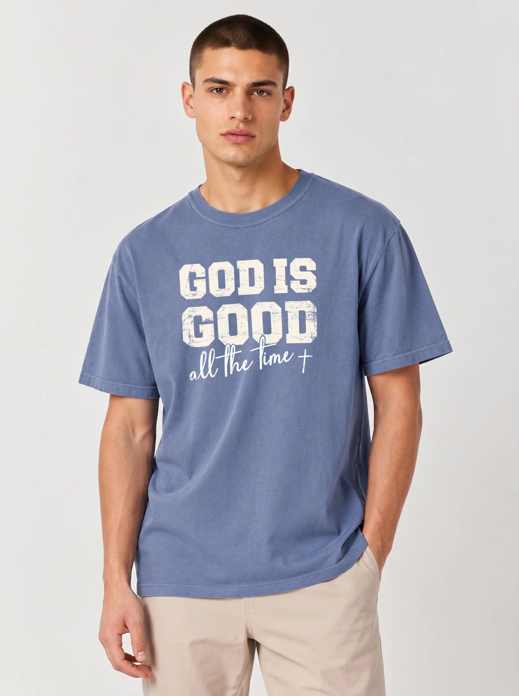 A man wearing a slate blue t-shirt with a white graphic print that reads 'GOD IS GOOD all the time +', paired with beige pants, against a light gray studio background.