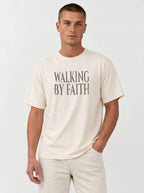Person wearing a cream T-shirt with distressed gray text reading “Walking By Faith.”