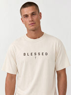 A man wearing a cream-colored crewneck t-shirt with the word 'BLESSED' and a small cross printed in black on the chest, set against a plain light gray background.