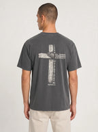 A man viewed from the back wearing a dark gray washed graphic t-shirt with a distressed biege graphic of a cross with the text 'I CAN DO ALL THINGS THROUGH CHRIST WHO STRENGTHENS ME' from Philippians 4:13, paired with light beige pants, against a plain light gray studio background.