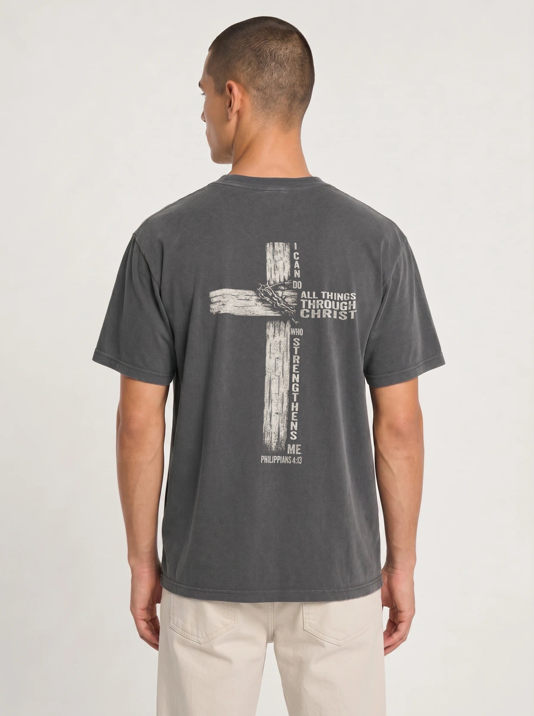 A man viewed from the back wearing a dark gray washed graphic t-shirt with a distressed biege graphic of a cross with the text 'I CAN DO ALL THINGS THROUGH CHRIST WHO STRENGTHENS ME' from Philippians 4:13, paired with light beige pants, against a plain light gray studio background.