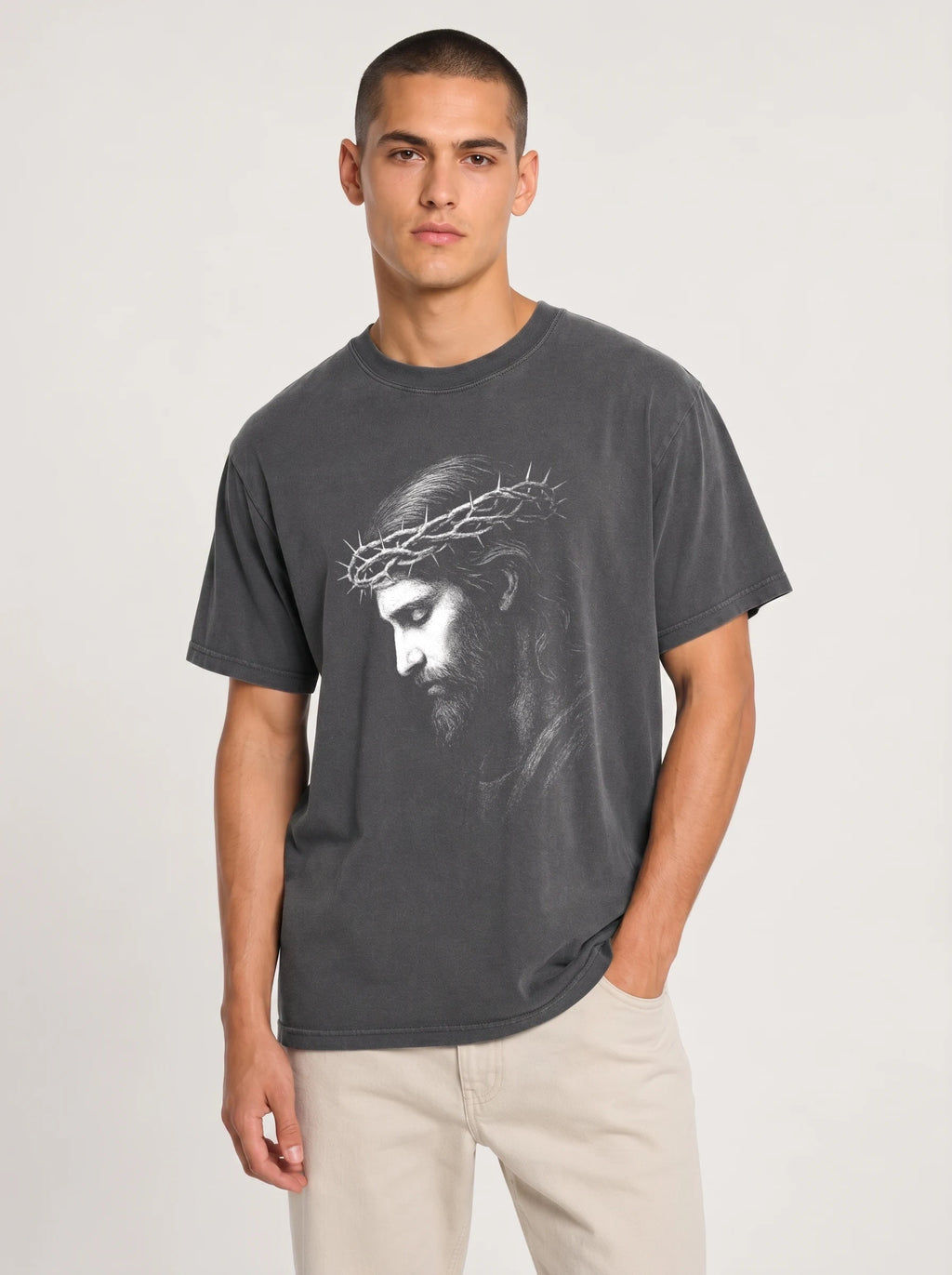 A man wearing a dark gray crewneck t-shirt with a white graphic print of Jesus' face wearing a crown of thorns, paired with light beige pants, against a light gray studio background.