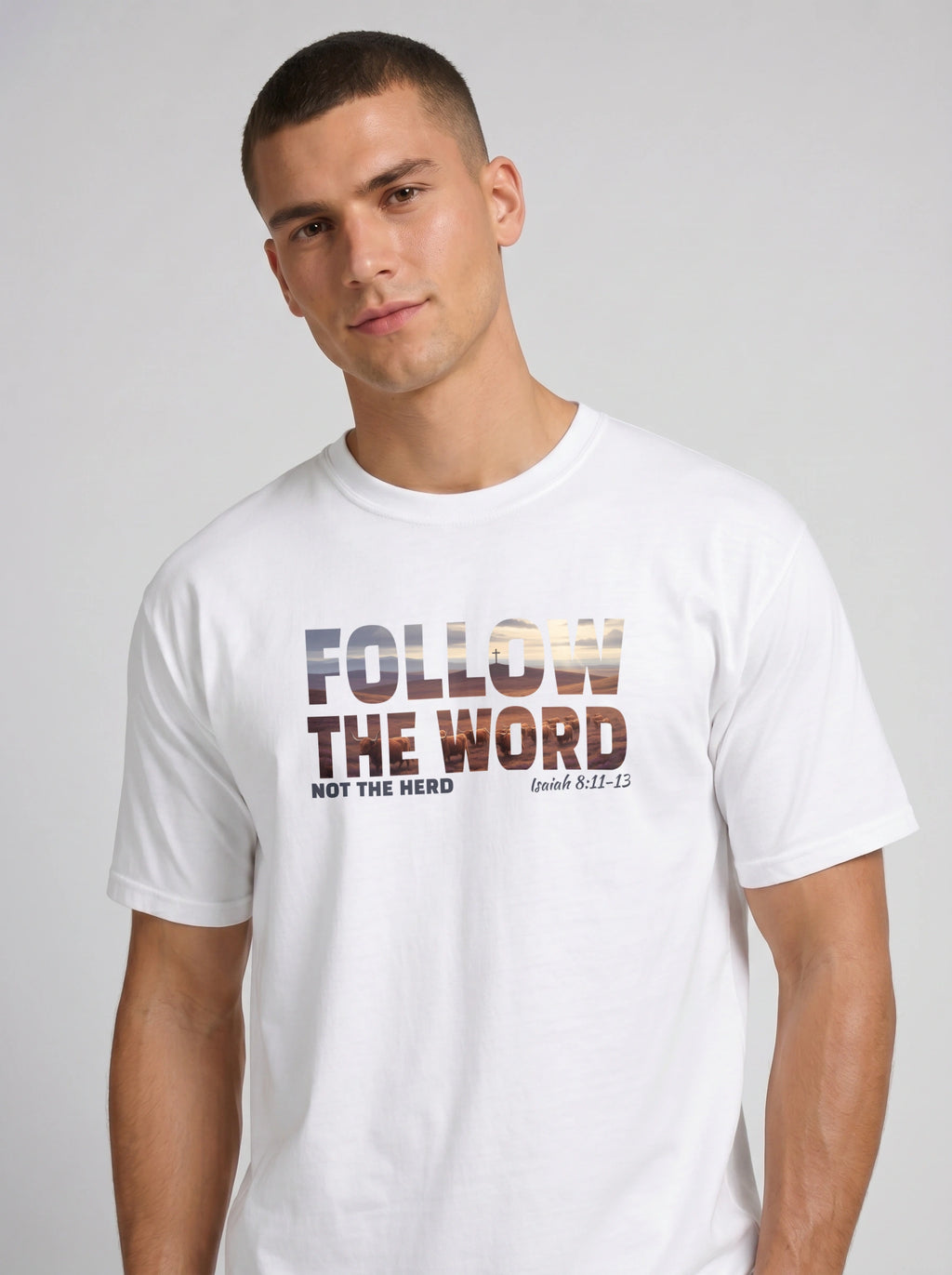 A man wearing a white crew-neck t-shirt with a graphic print that reads 'FOLLOW THE WORD NOT THE HERD' and 'Isaiah 8:11-13' in a textured, multi-toned font' in a textured, multi-toned font. The photo is taken against a neutral light gray background.