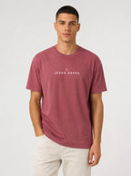 A man wearing a dusty rose short-sleeve t-shirt with the white printed text 'JESUS SAVES' and a small cross on the chest, paired with off-white pants, against a light gray studio background.