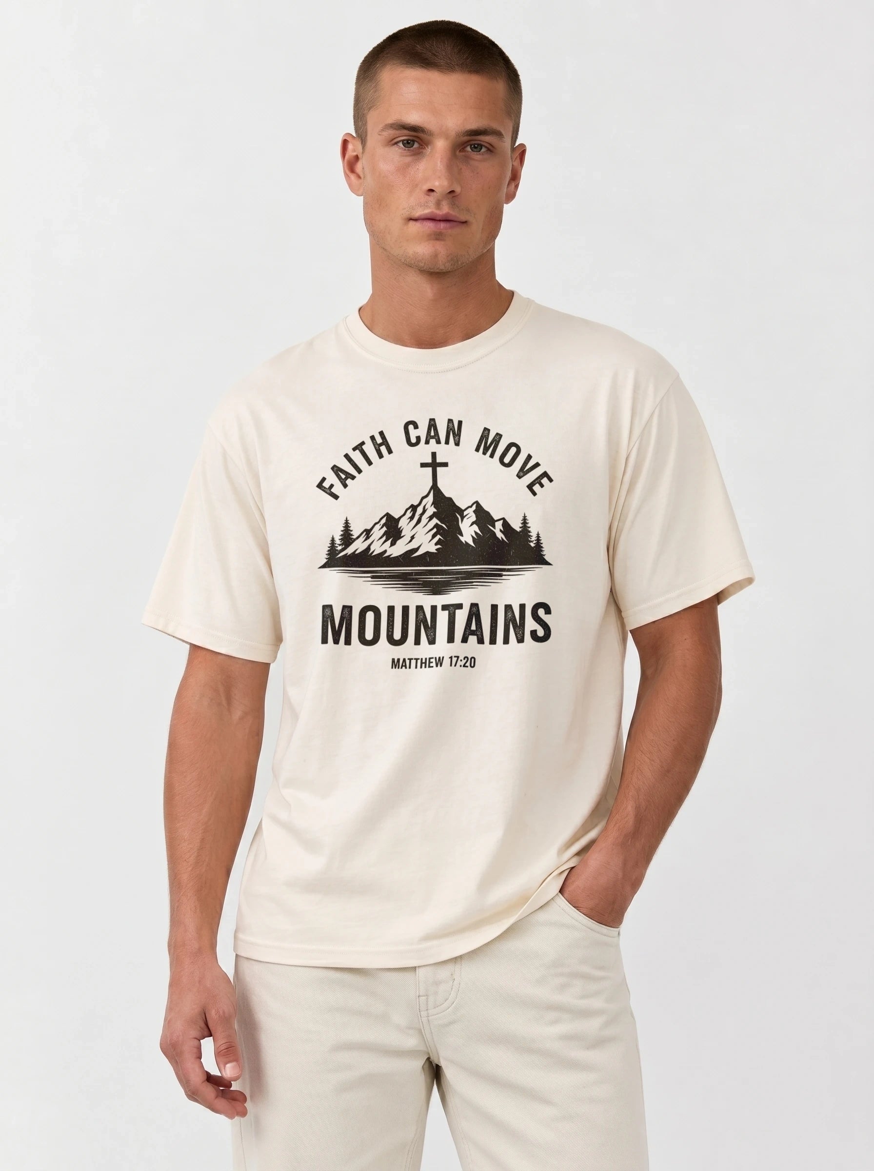A man wearing a cream-colored t-shirt with a black graphic print that reads 'FAITH CAN MOVE MOUNTAINS MATTHEW 17:20' with a mountain and cross illustration, paired with light beige pants, against a plain light gray background.