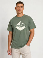 A man wearing an olive green crewneck t-shirt with a white mountain graphic and the text 'BE STILL' on the chest, paired with beige pants, against a light gray studio background.