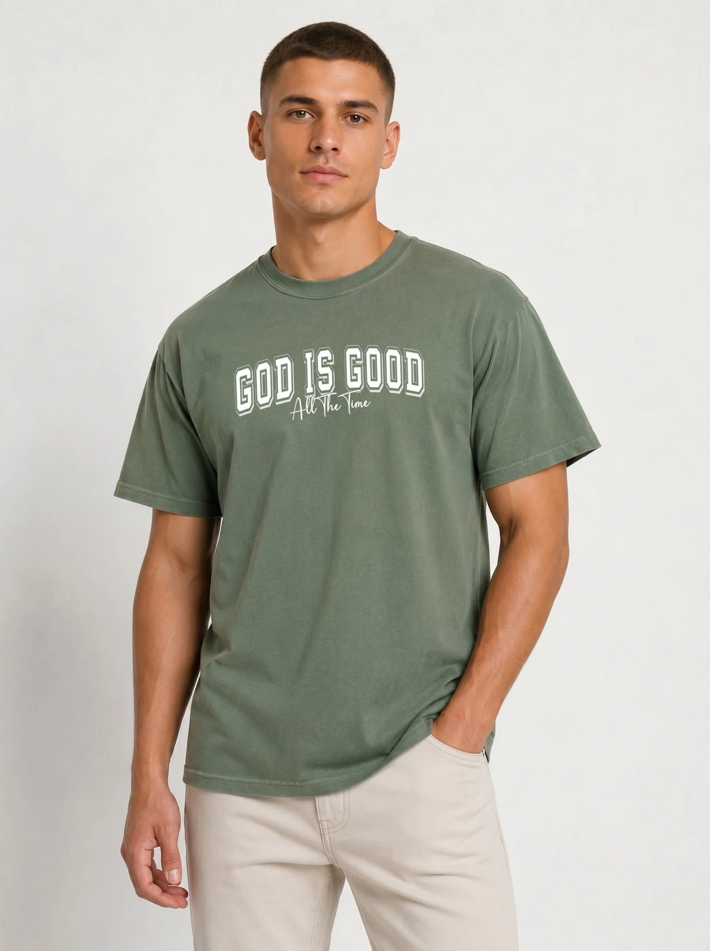 A man wearing a muted sage green t-shirt with 'GOD IS GOOD' printed in white across the chest, paired with light beige trousers, against a plain light gray background.