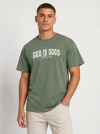 A man wearing a muted sage green t-shirt with 'GOD IS GOOD' printed in white across the chest, paired with light beige trousers, against a plain light gray background.
