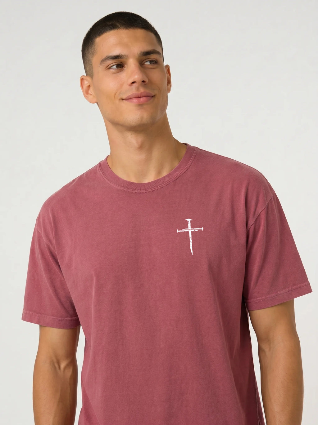 A man wearing a dusty rose short-sleeve t-shirt with a white cross graphic print the chest, set against a plain light gray studio background.