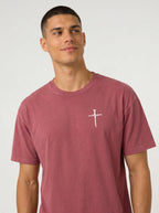A man wearing a dusty rose short-sleeve t-shirt with a white cross graphic print the chest, set against a plain light gray studio background.