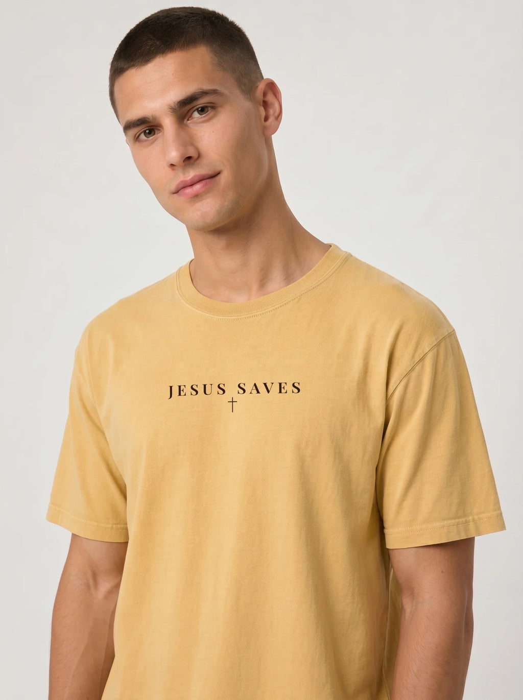 Young man modeling a mustard yellow T-shirt featuring black “JESUS SAVES” text with a small cross below, posed against a plain light background.