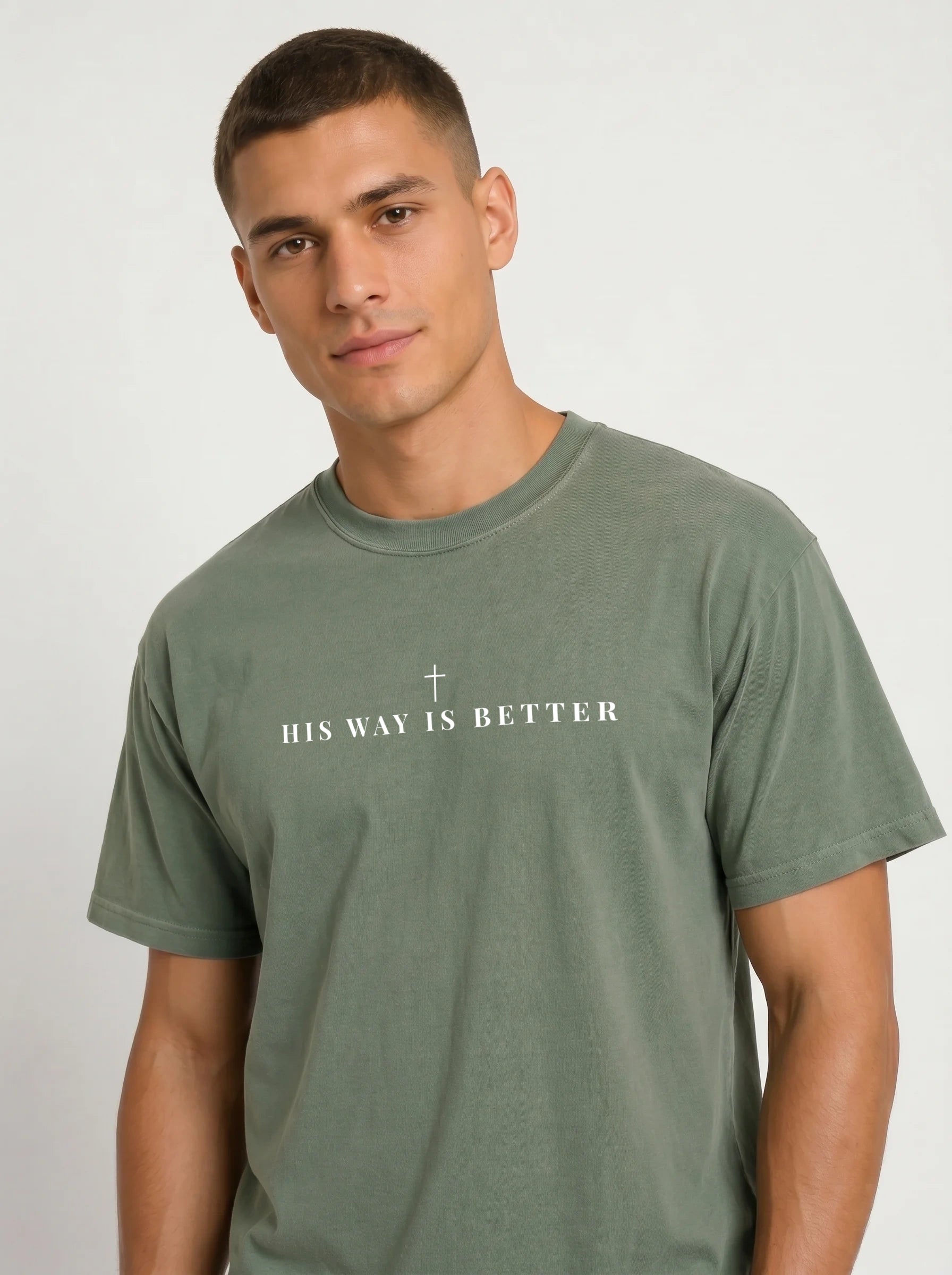 A man wearing a muted sage green crewneck t-shirt with a white graphic text print on the chest that reads 'HIS WAY IS BETTER' and a small cross.