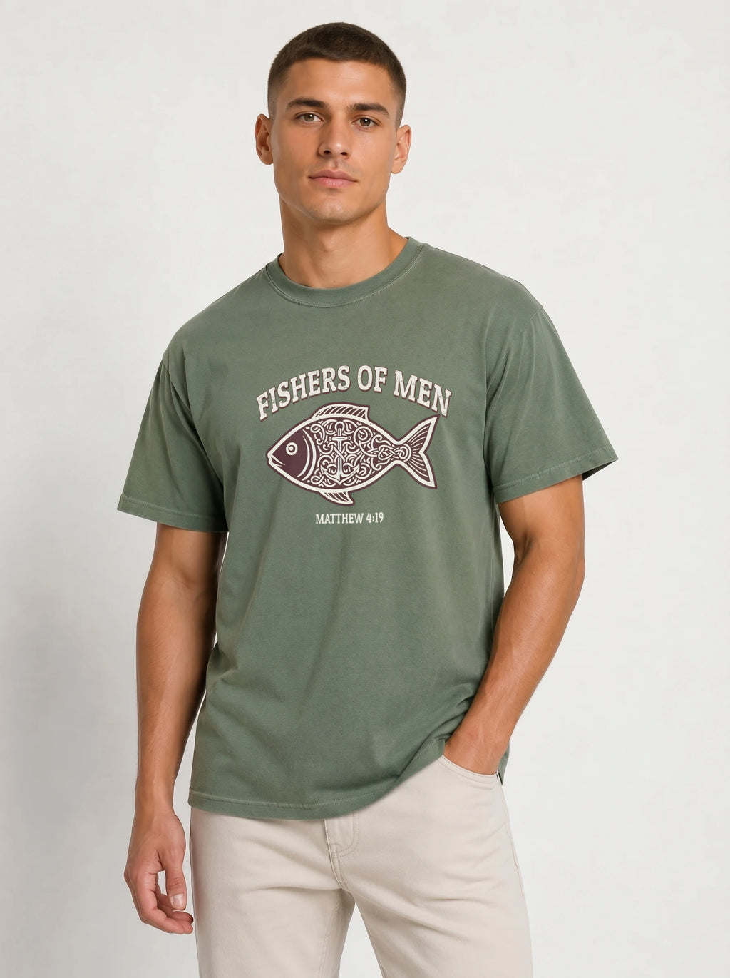 A man wearing an olive green t-shirt with a 'FISHERS OF MEN' graphic on the chest, paired with beige pants, standing against a light gray studio background.