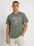 A man wearing an olive green t-shirt with a 'FISHERS OF MEN' graphic on the chest, paired with beige pants, standing against a light gray studio background.