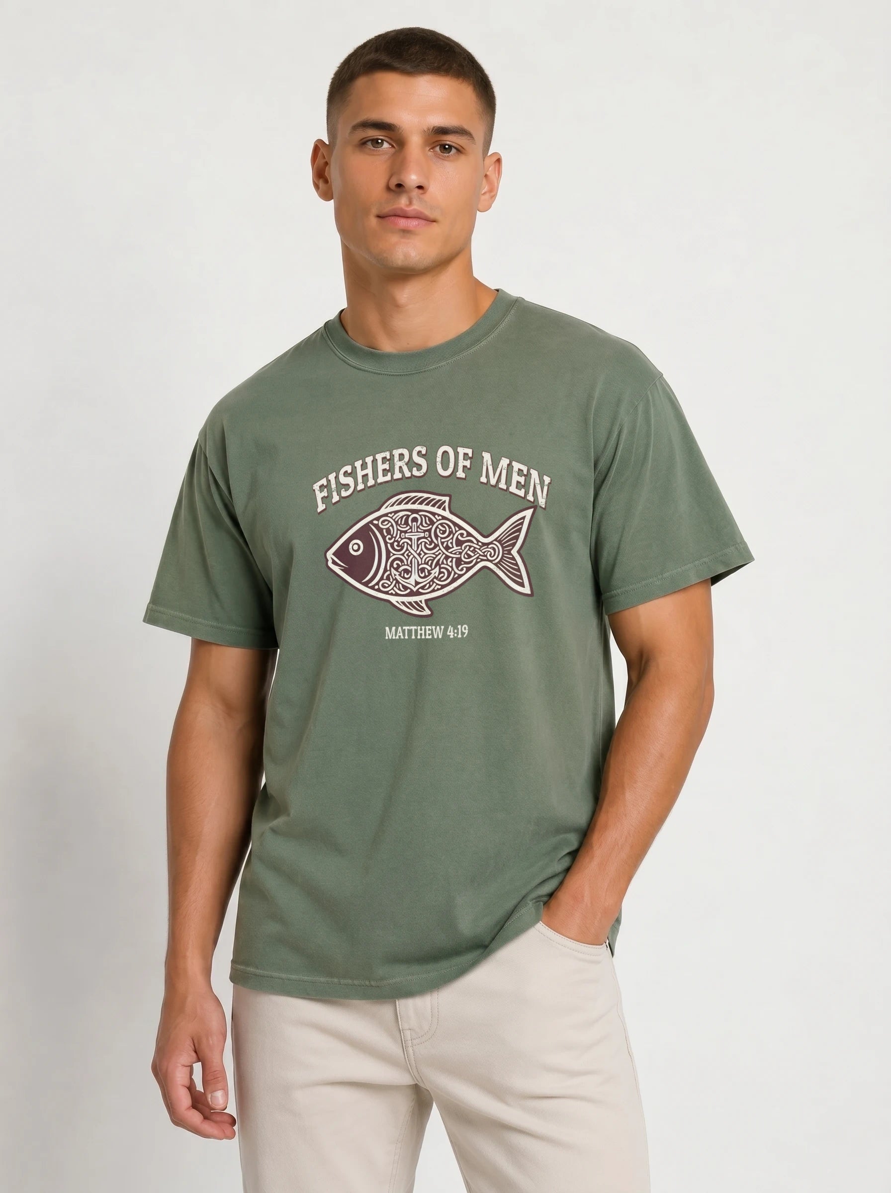A man wearing an olive green t-shirt with a 'FISHERS OF MEN' graphic on the chest, paired with beige pants, standing against a light gray studio background.